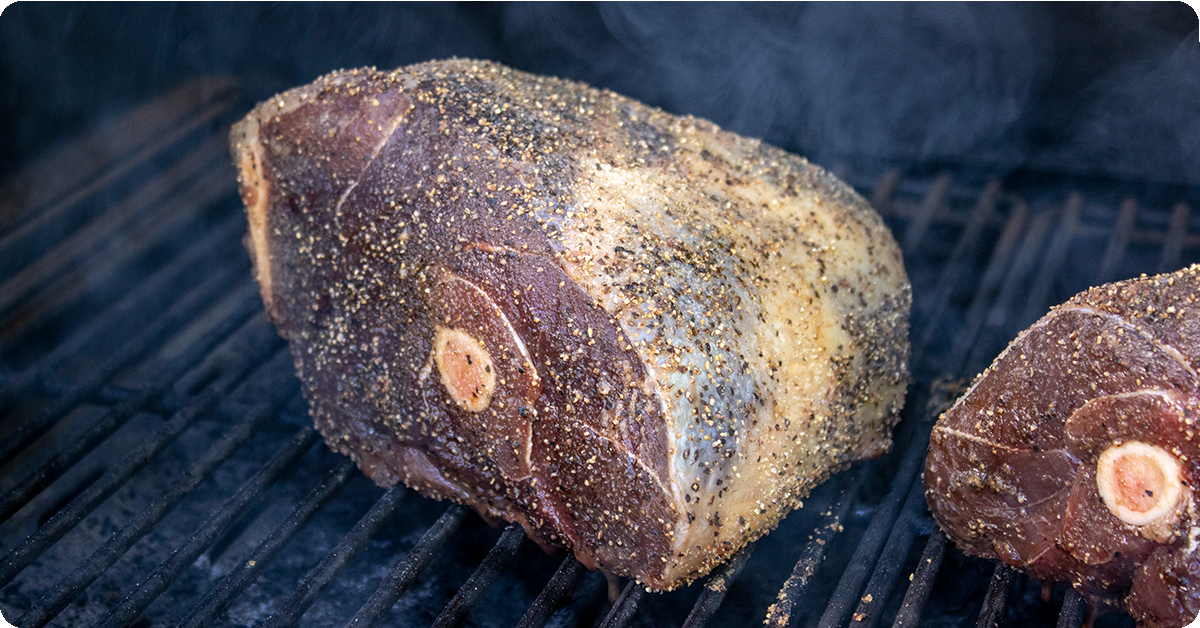 Two bone-in legs of lamb being smoked on a grill.