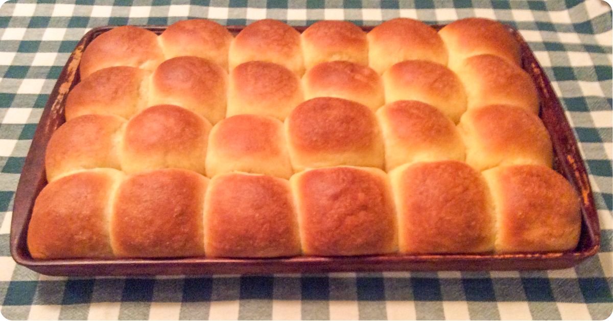 A tray of perfectly browned homemade rolls on a checkered country table cloth.