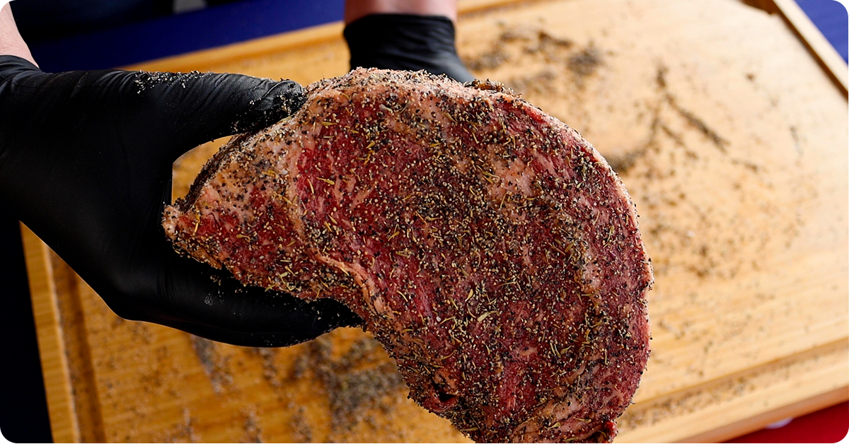 A pair of gloved hands raise up a a well-seasoned prime rib roast.